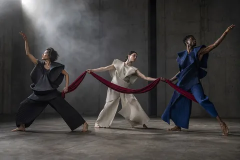 Three dancers sharing a flowing red ribbon in a gray studio